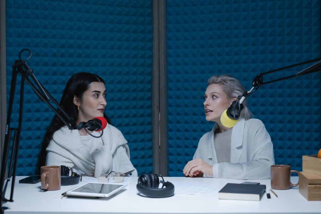 Two women engaged in conversation, recording podcast in studio with microphones and headphones.